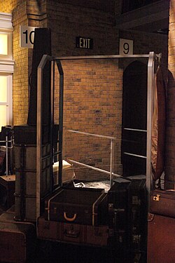 Photograph of a hotel-style metal-framed luggage trolley surrounded by old-style suitcases. In the background is what appears to be a solid brick wall. with platform number sign „10“ on the left, and „9“ on the right. The trolley contains a human-sized semi-transparent mirror that combines the foreground and background.