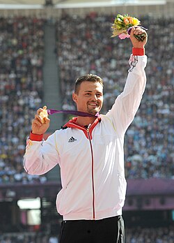 Oq sport kiyimida, guldasta ushlab turgan va boshqa qoʻlida oltin medalni koʻtarib turgan oq tanli erkak.