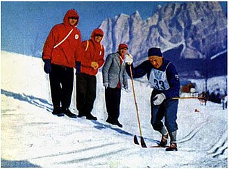A man in blue clothes with a number „33“ cross-country skiing in front of three other men.