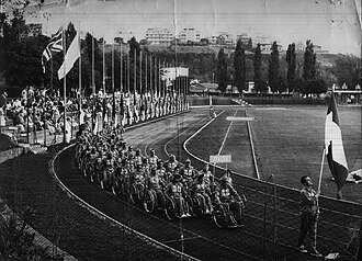 A black and white photo of a large track and field stadium with spectator stands and flags flying on the left hand side and trees in the background. A large procession of people in wheelchairs goes round the track with a sign being held at the front reading „Italia“. The team is led by a walking man carrying the Italian flag.