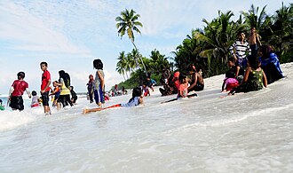 Qoramtir terili oʻgʻil va qizlar sohil toʻlqinlari orasida hoʻl kiyimda tik turgan, oʻtirgan va yotgan surat. Orqa fonda bir nechta palma daraxtlari koʻrinadi. Bolalar orasida niqobli ayollar ham bor.