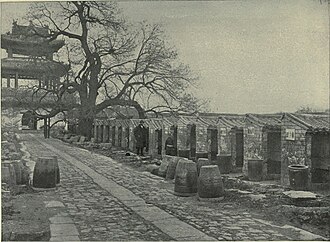 A black-and-white picture of a stone-paved alley going from bottom right to top left leading to a three-roofed gate and bordered on the right by a line up of small roofed cubicles open on one side.
