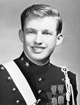 A black-and-white photograph of Trump as a teenager, smiling, wearing a dark pseudo-military uniform with three ribbons and a white shoulder strap